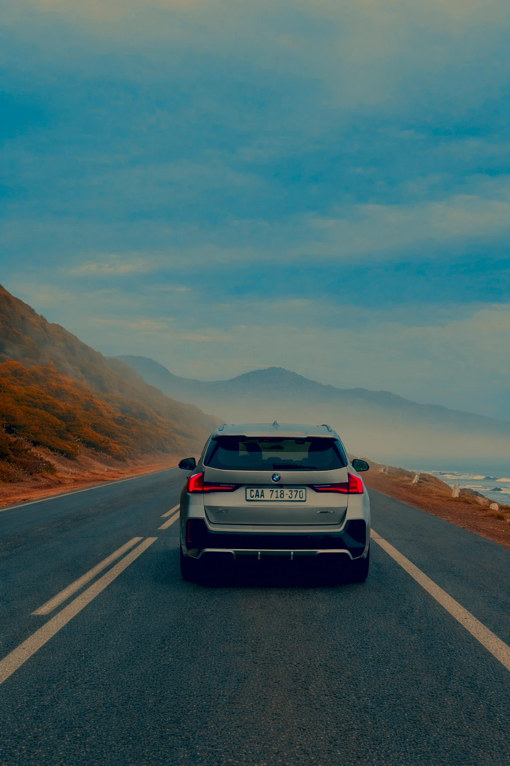Silberner SUV von hinten auf einer Küstenstraße mit Nebel und Bergen im Hintergrund